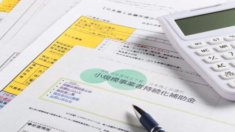 小規模事業者持続化補助金の申請書類と電卓・ボールペンの写真。中小企業の資金調達や補助金活用のイメージ。