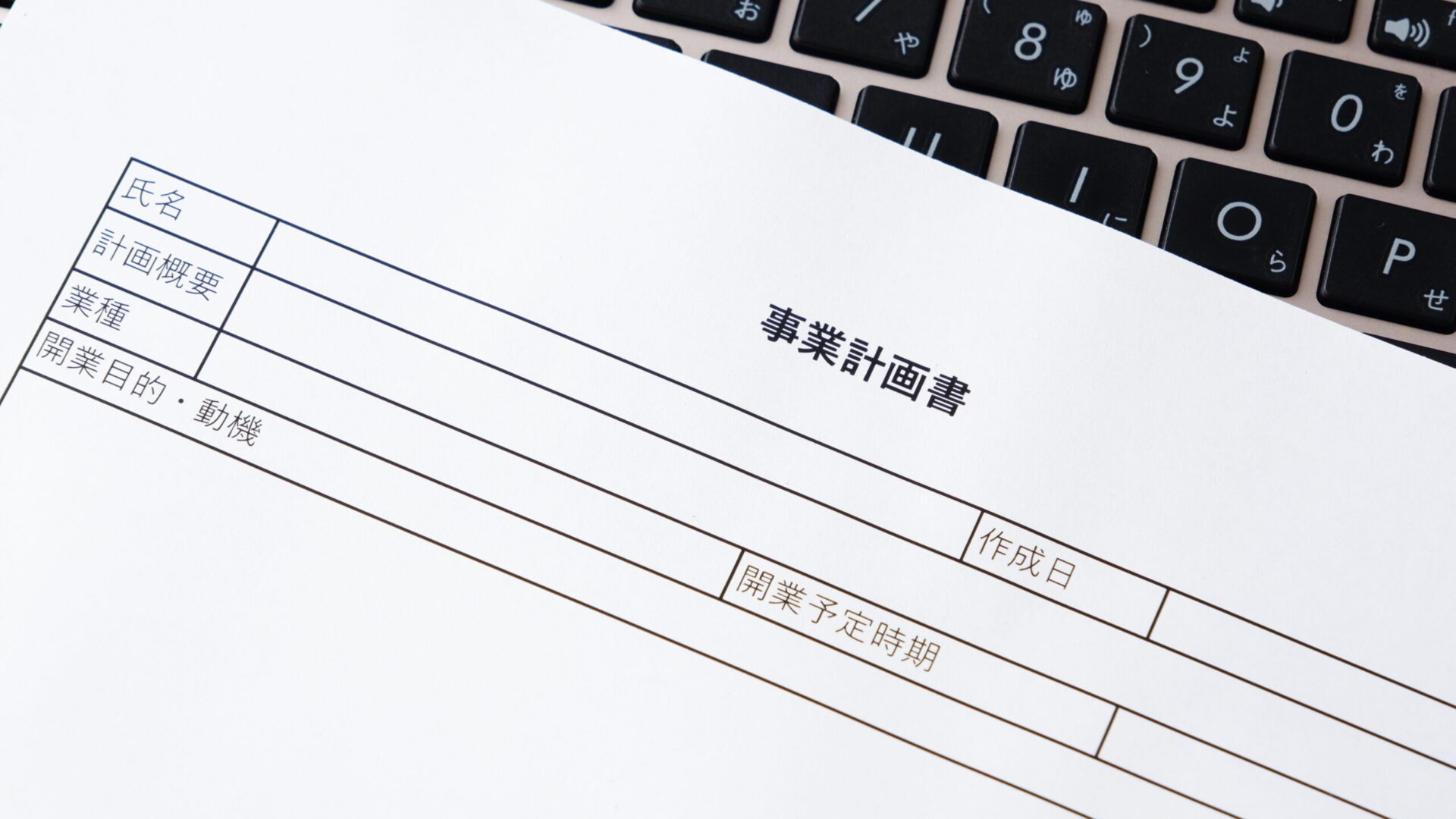 ノートパソコンのキーボードの上に置かれた事業計画書の用紙。補助金申請や開業計画の作成をイメージした写真。