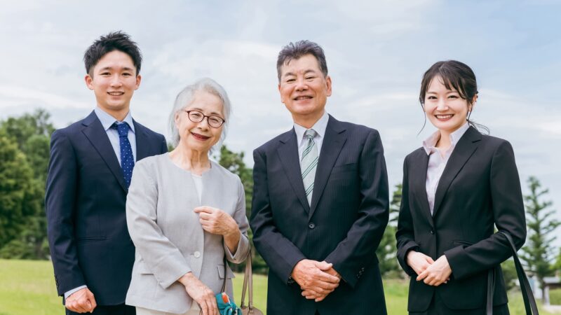 スーツ姿の家族が屋外で笑顔を見せている。世代を超えて中小企業経営を支える家族のイメージ。