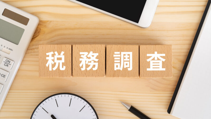 木製ブロックに「税務調査」と書かれた文字が並び、机の上に電卓・時計・スマートフォン・ノート・ペンが置かれている