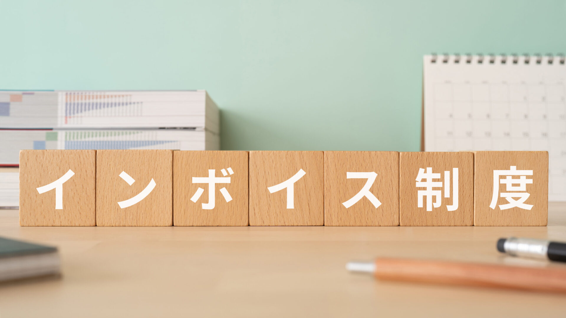 「インボイス制度」と書かれた木製ブロックが机上に横一列に並び、背景にカレンダーと資料、手前にペンが置かれている。