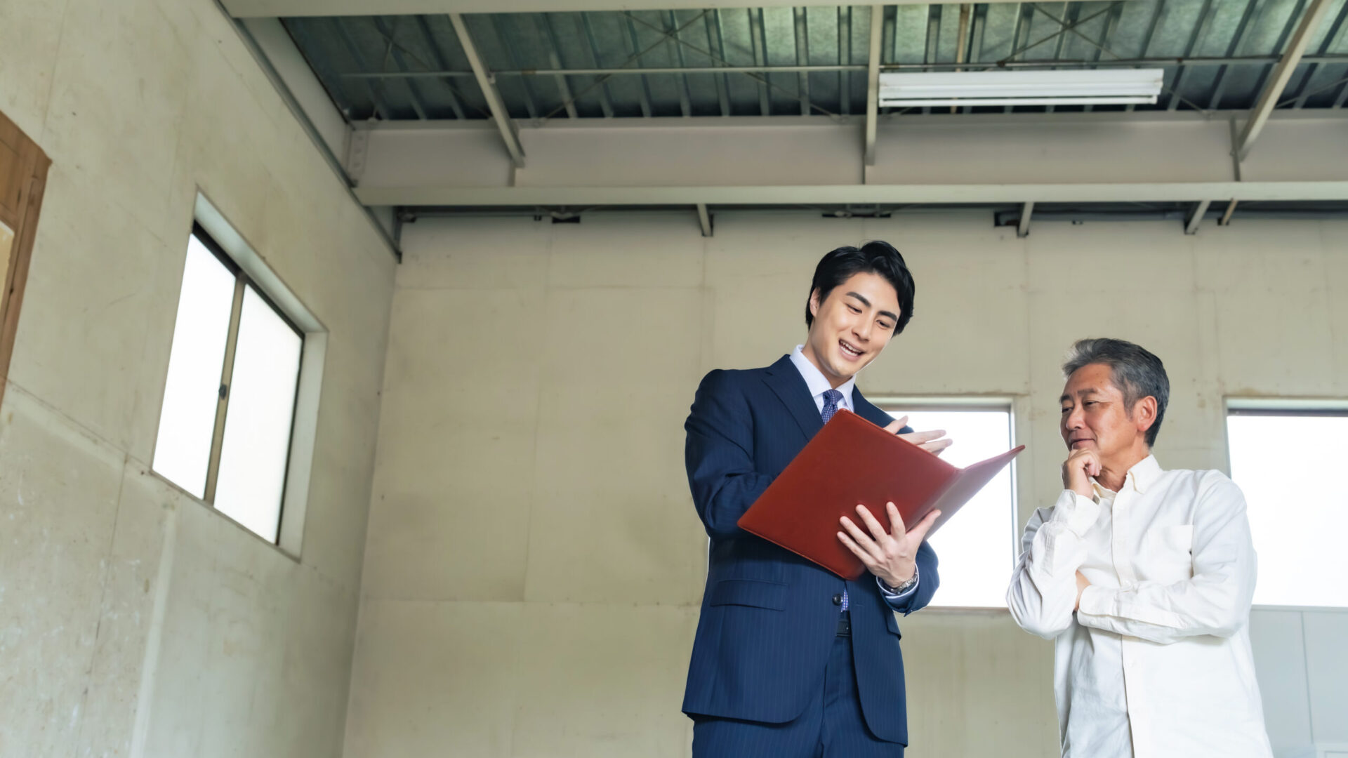 改装前の事務所で、スーツ姿の担当者が図面の入ったファイルを見せながら、中年男性の経営者に工事内容を説明している様子