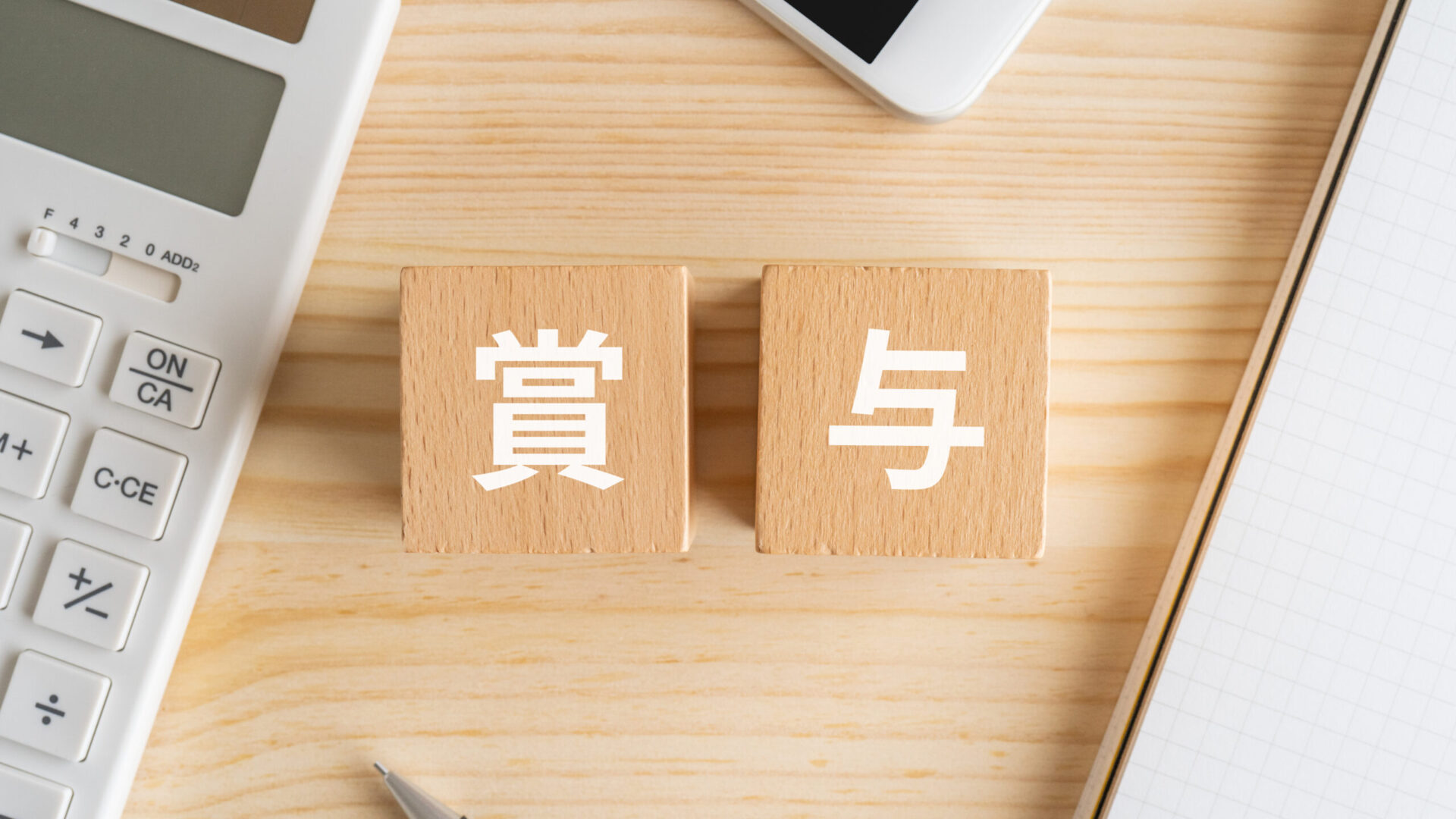 木製ブロックに「賞与」と書かれた文字と、電卓・スマートフォン・ノートが並ぶデスク。年末の賞与計算や役員報酬設定をイメージしたビジネスシーン。