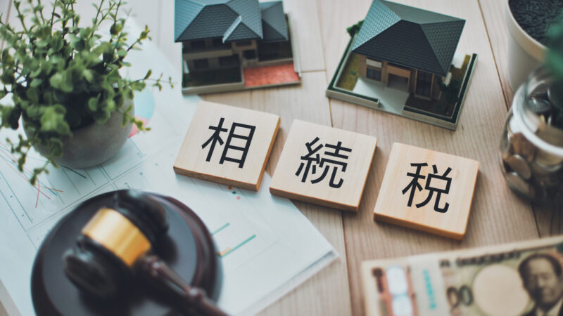 家や不動産の模型と相続税と書かれた木のブロック、判決文のハンコに見立てた木槌と日本円が並ぶ相続税のイメージ