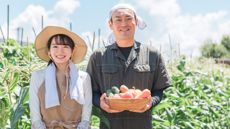 畑で収穫した野菜を手に笑顔で並ぶ農業従事者2人の写真