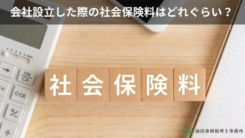 会社設立時の社会保険料について解説するバナー画像。木目の机の上に「社会保険料」と書かれた積み木、電卓、ペン、ノートが配置されている。