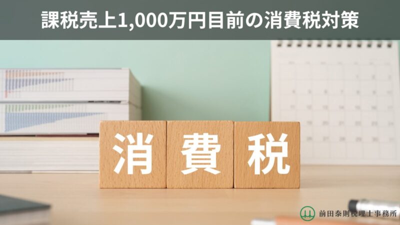 「課税売上1,000万円目前の消費税対策」という見出しと、木製ブロックで「消費税」と表現した画像。背景には本棚とカレンダーがあり、右下に前田泰則税理士事務所のロゴと事務所名が入っている。